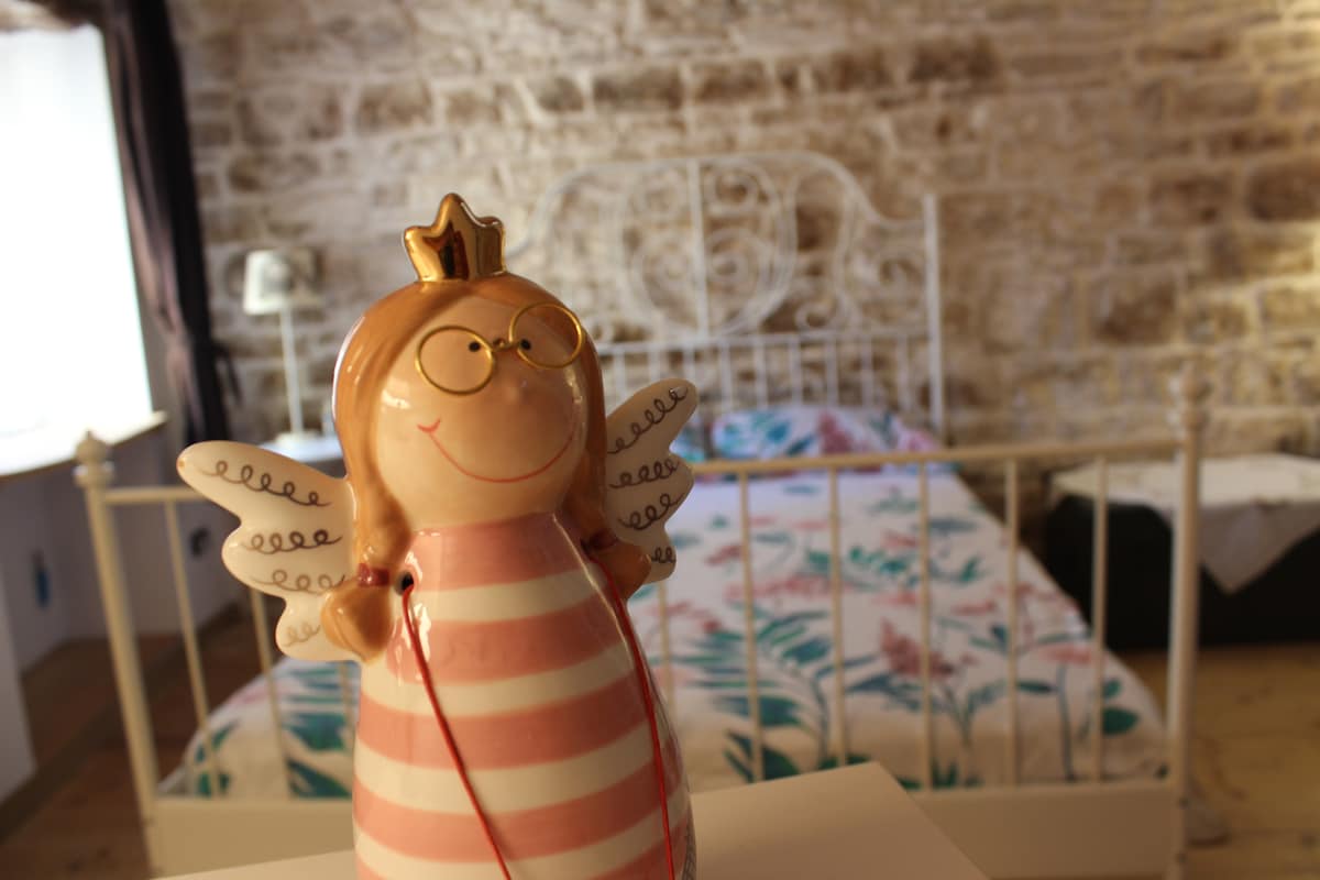 A decorative figure with a crown and wings is positioned in the foreground. In the background, a double bed is visible with a floral-patterned blanket, set against a rustic stone wall. Soft natural light filters through a nearby window.