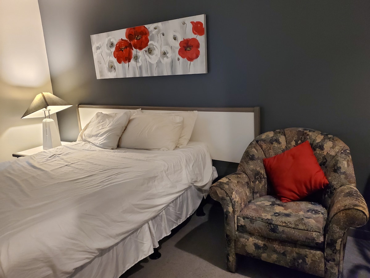 A sleeping area features a spacious bed with white bedding, complemented by a floral artwork with red and white tones above. A patterned armchair with a red accent pillow sits nearby, alongside a lamp on a bedside table, providing soft illumination.