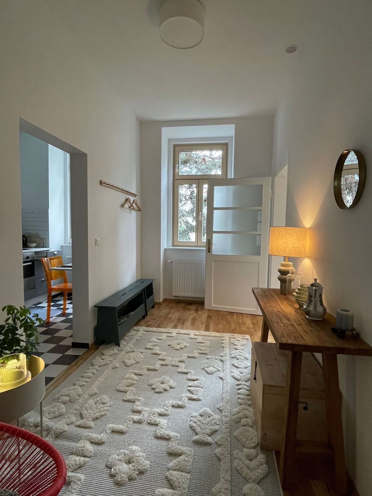 A bright entryway is showcased, featuring a patterned area rug and wooden furniture. Natural light filters through a window, illuminating a console table adorned with decorative items. A wall-mounted coat rack stands beside an open door leading to the kitchen, enhancing the inviting atmosphere.