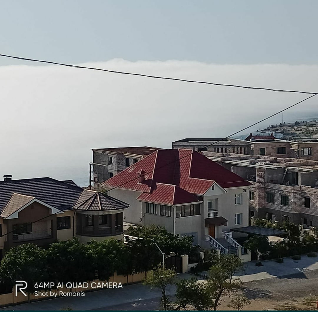 A vibrant red roof contrasts with the surrounding buildings, set against a misty backdrop. The neighborhood features a mix of architectural styles, with greenery visible at the forefront. The image captures a serene atmosphere with a hint of the ocean beyond the structures.