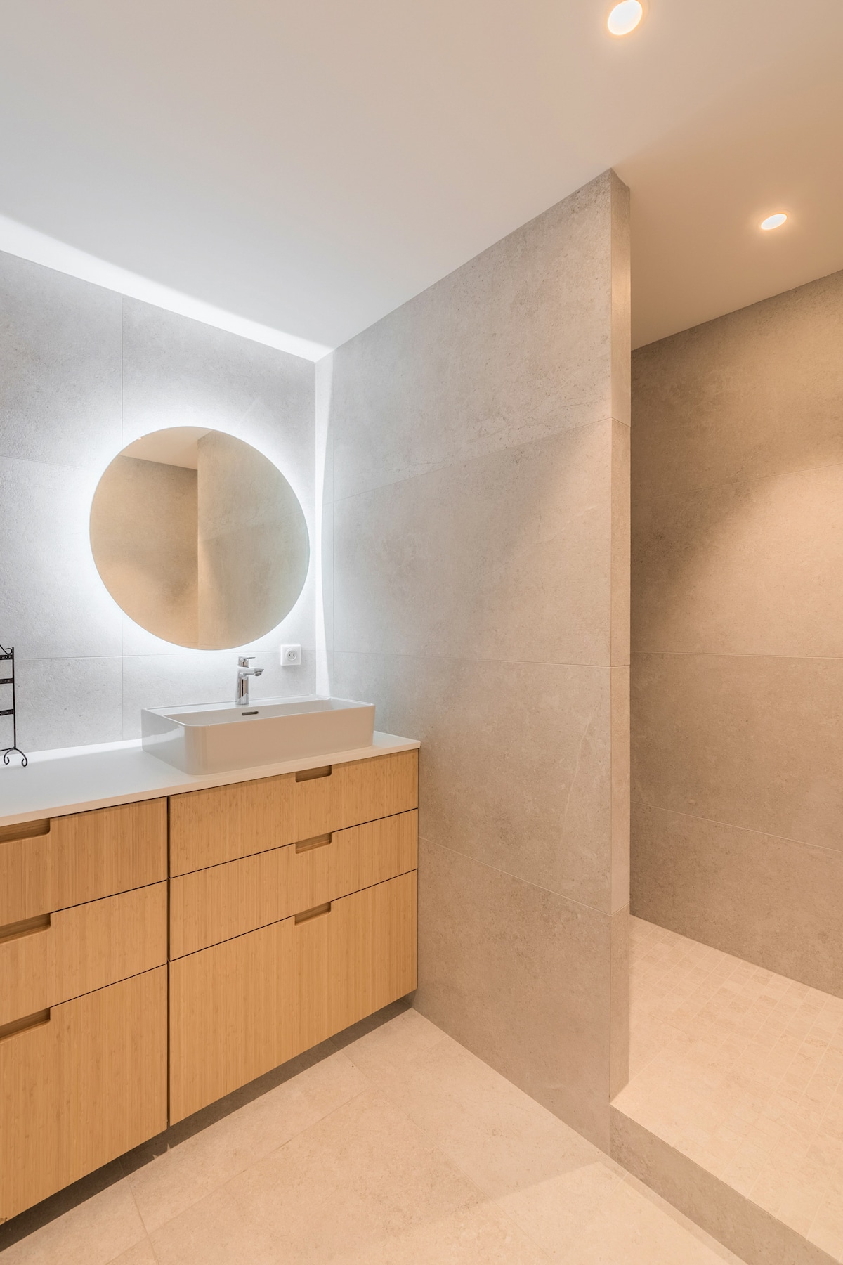 A modern bathroom features soft lighting illuminating a round mirror above a minimalist sink. Textured gray tiles line the walls, while a spacious shower area is visible, showcasing seamless design and elegant fixtures.