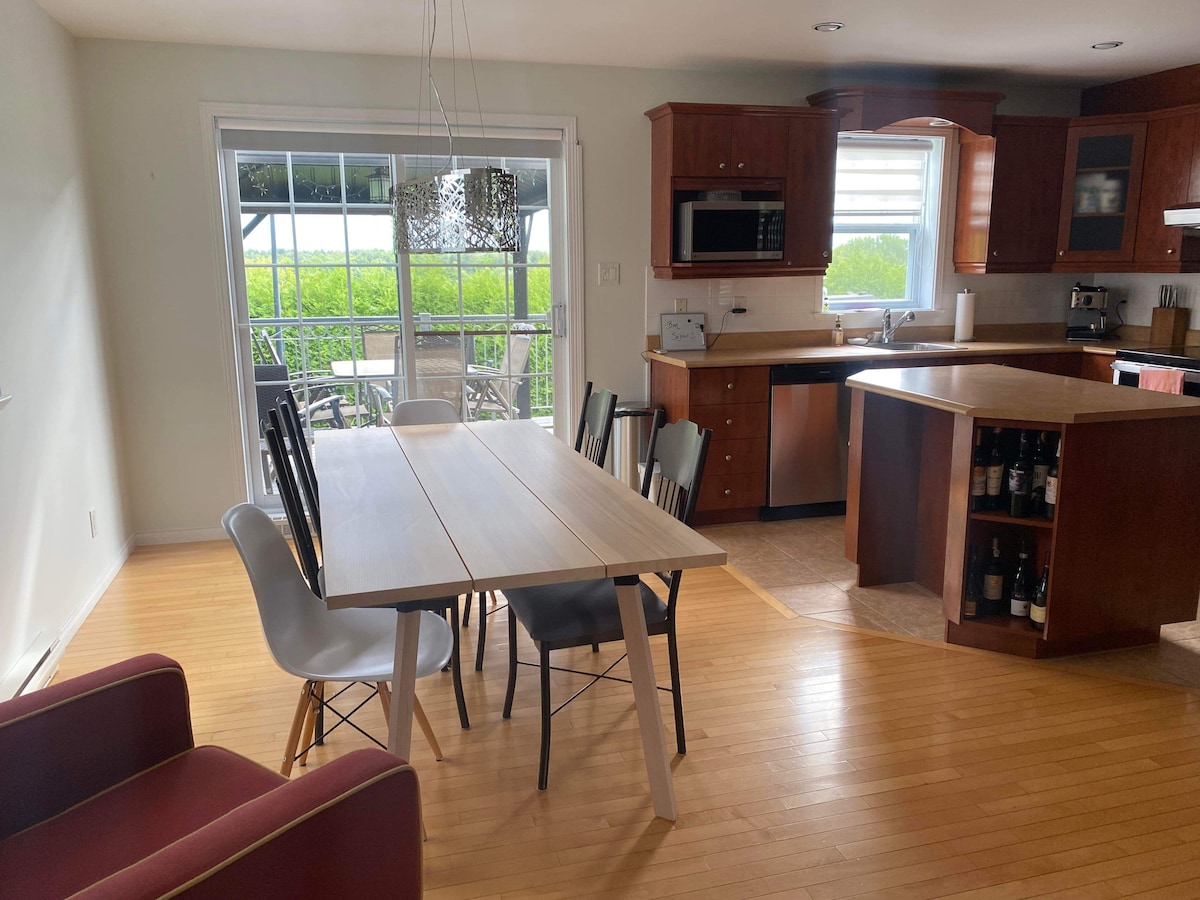 A dining area features a long table surrounded by various chairs, including modern and classic styles. Light wood cabinetry and surfaces are complemented by a window that overlooks a green outdoor space. An open kitchen layout is visible, showcasing appliances and a central island.