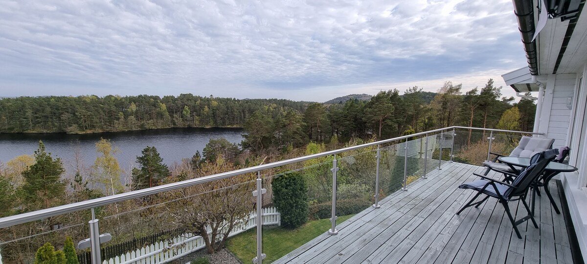 A spacious deck offers a peaceful view of the surrounding trees and a serene lake. Two lounge chairs are positioned for relaxation, while a glass railing enhances the open feel of the outdoor space.