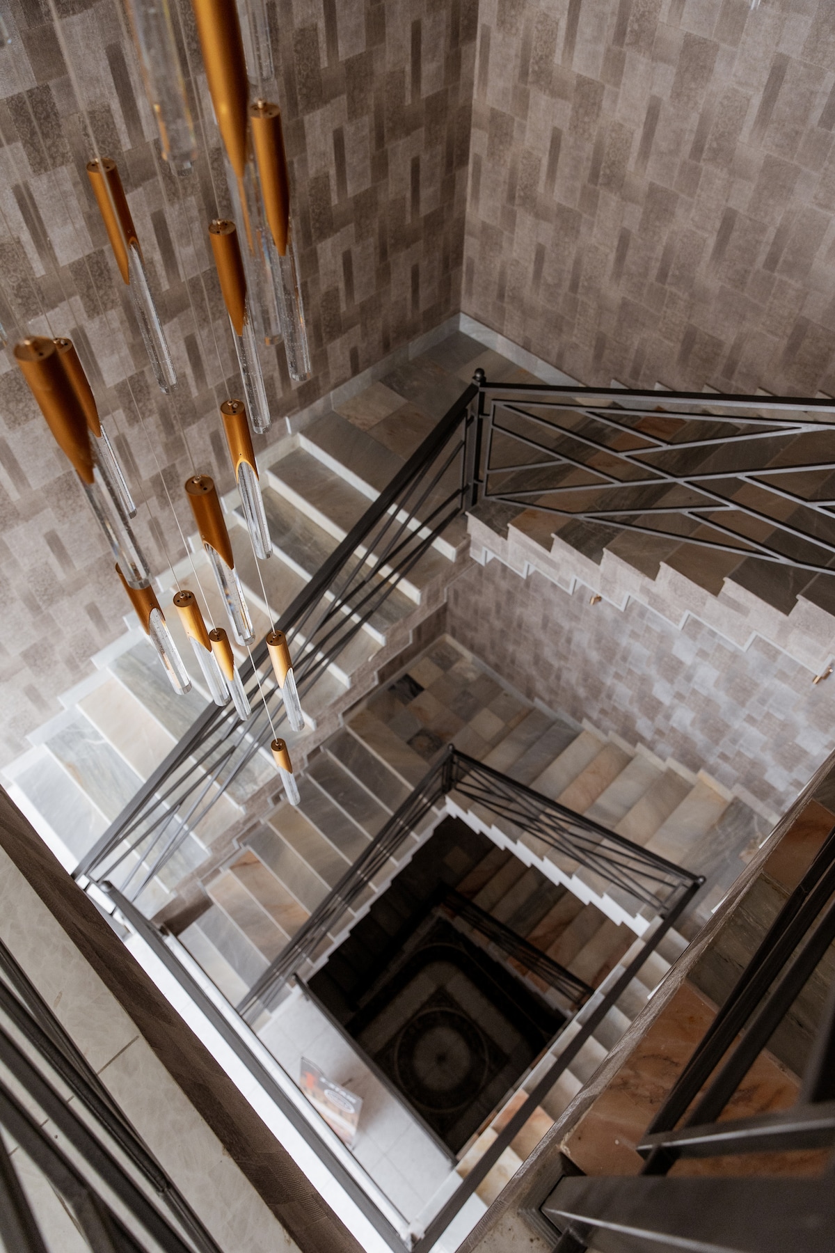 A multi-level staircase features a sleek design with stone steps and metal railings. A series of elongated light fixtures hang gracefully from the ceiling, casting soft illumination. The textured wall tiles add depth and dimension to the space, enhancing its modern aesthetic.