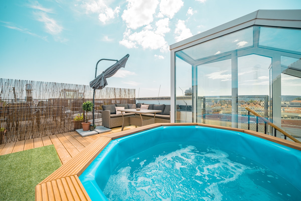 A well-appointed rooftop terrace features a circular jacuzzi surrounded by wooden decking. Comfortable outdoor seating is offered beneath a large umbrella, and a glass enclosure provides protection while allowing for a scenic view. Decorative plants enhance the inviting atmosphere of the space.