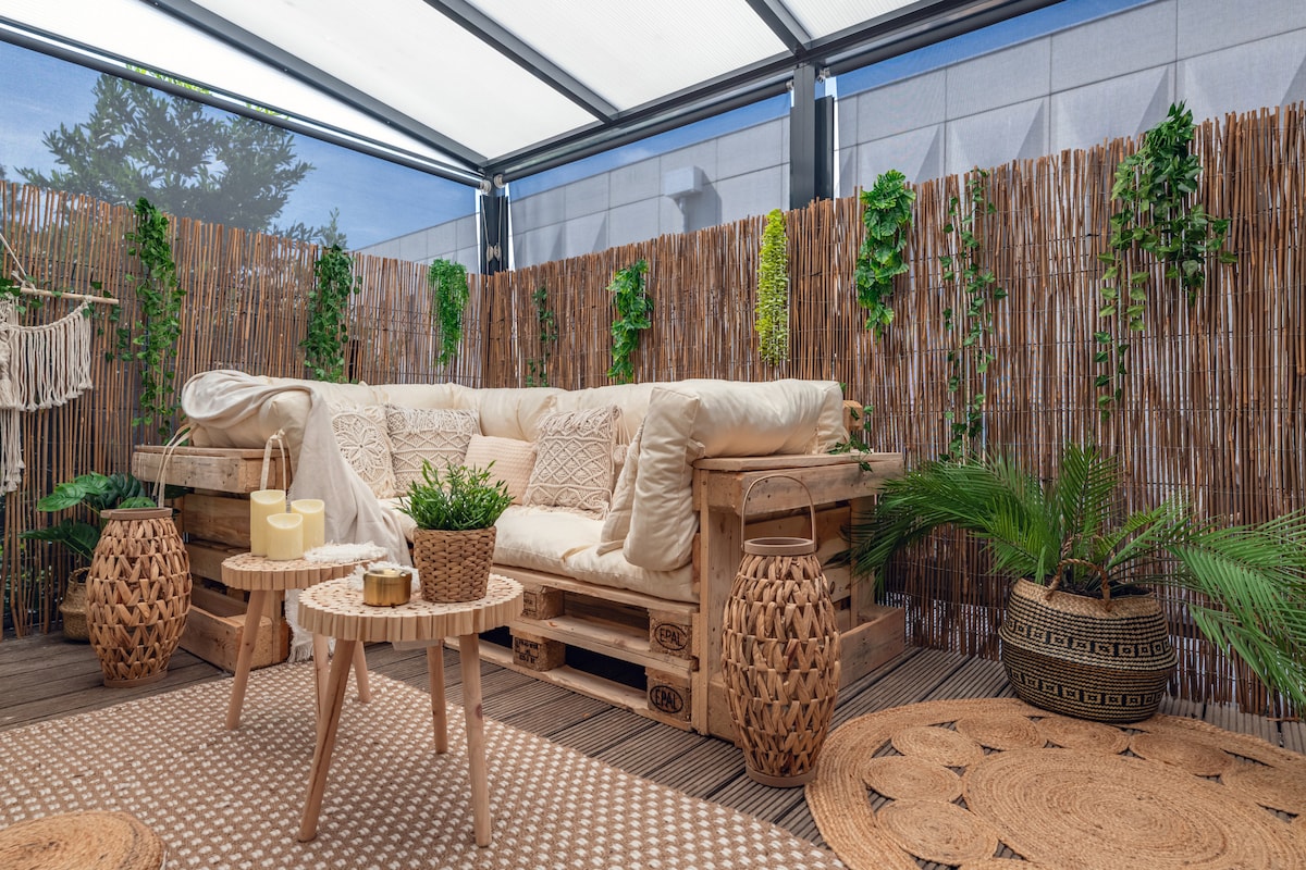 A cozy terrace area is presented, featuring a comfortable pallet sofa adorned with neutral-toned cushions. Several decorative plants enhance the space, accompanied by woven baskets and two small tables. A textured rug adds warmth to the wooden floor beneath a sheltered, transparent roof.