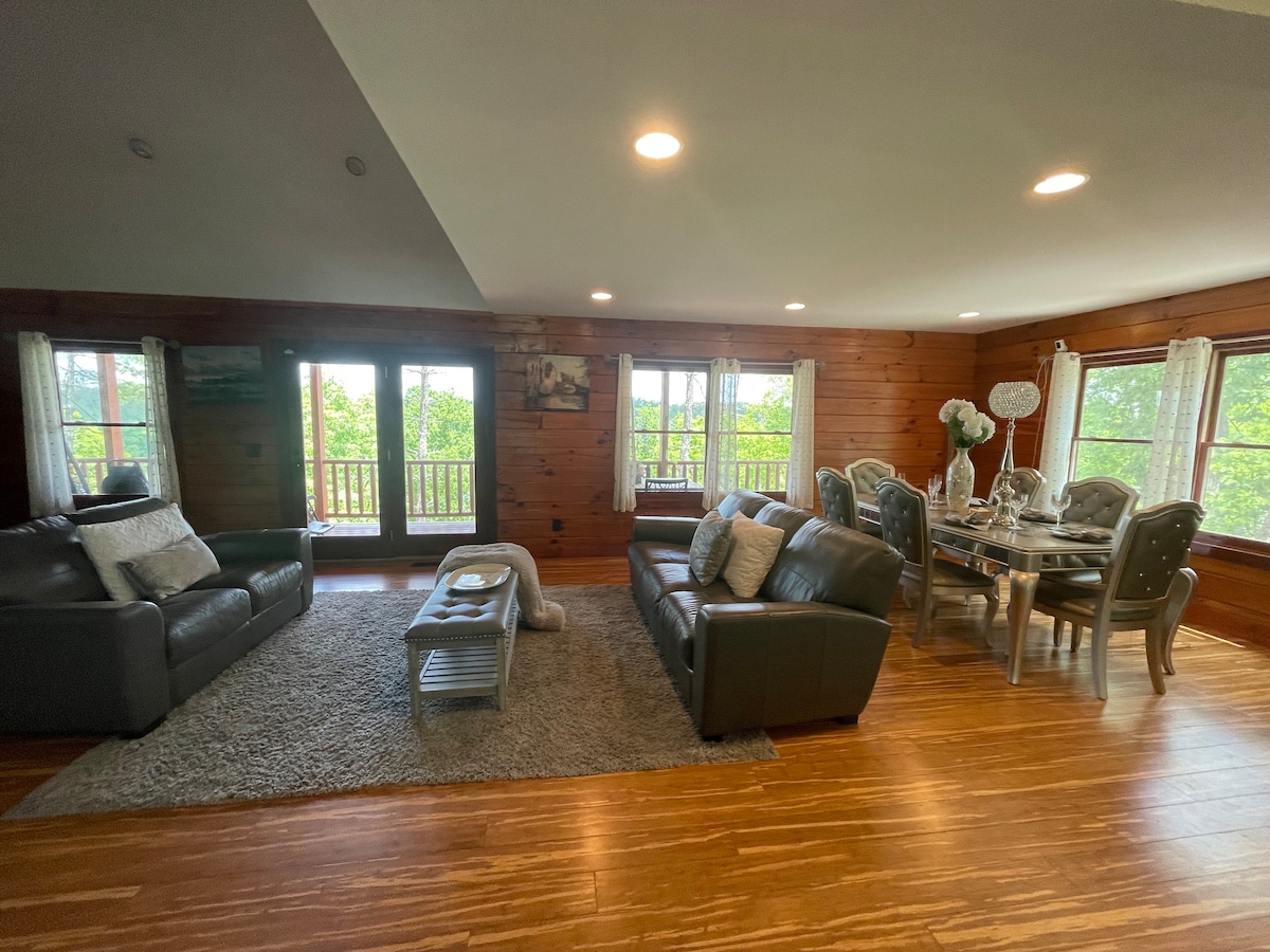 A spacious living area is highlighted by large windows that provide views of the outdoors. Comfortable leather sofas are arranged around a coffee table on a soft rug, while a dining table sits adjacent, accommodating multiple guests. Natural light fills the room, enhancing the wooden accents.