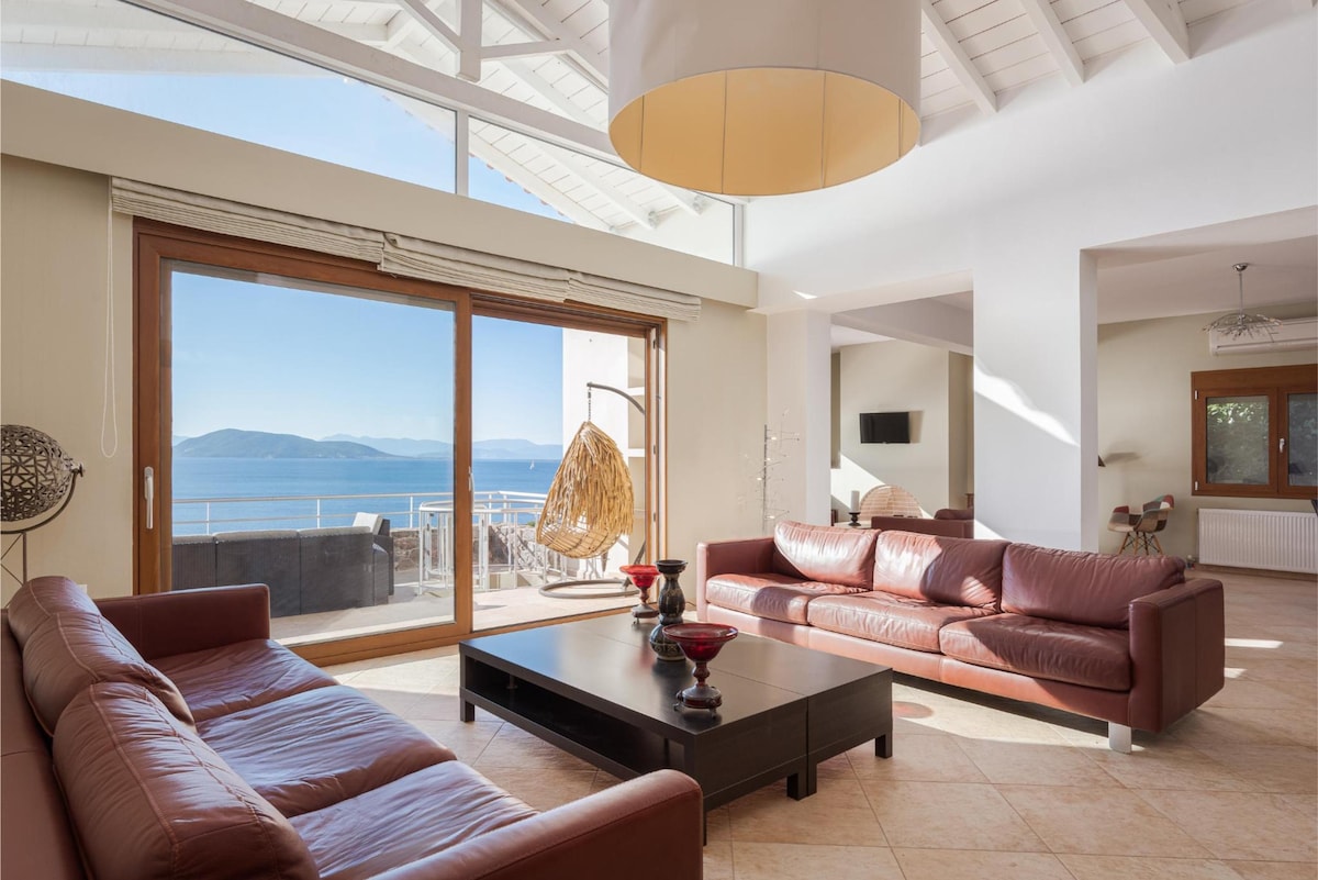 A spacious living area features large windows offering panoramic views of the sea and surrounding landscape. Comfortable leather sofas are arranged around a dark coffee table. Natural light fills the room, highlighting the high ceilings and tranquil atmosphere.