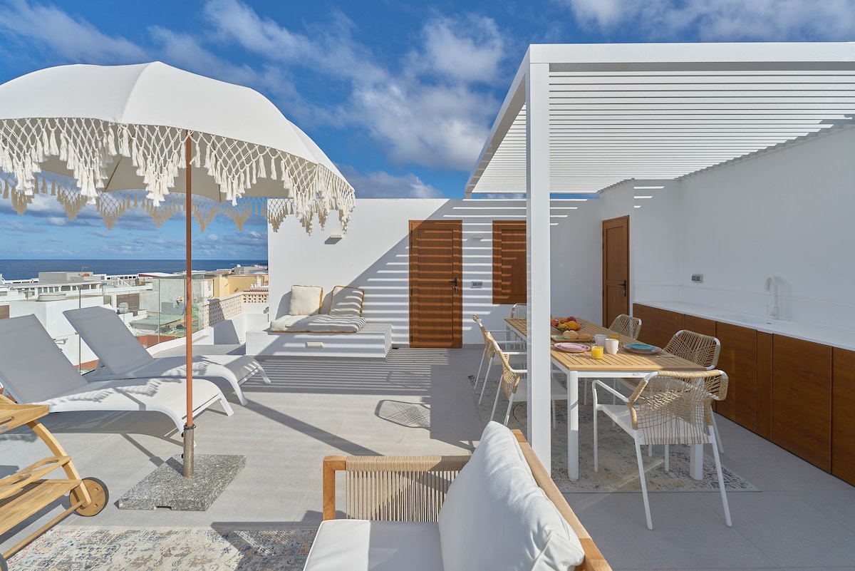 A spacious rooftop terrace is shown, featuring comfortable lounge chairs and a dining area. A stylish umbrella offers shade, while an outdoor kitchen is visible in the background. Clear blue skies and ocean views create a serene environment for outdoor gatherings.