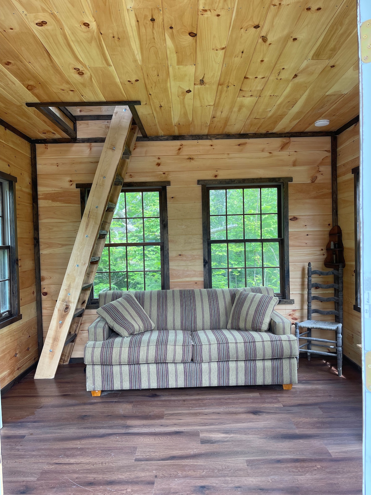 The interior features warm wood paneling with a striped couch positioned against the wall. A wooden ladder leads to a loft area above. Two windows are present, allowing natural light to brighten the space, which is complemented by dark flooring.