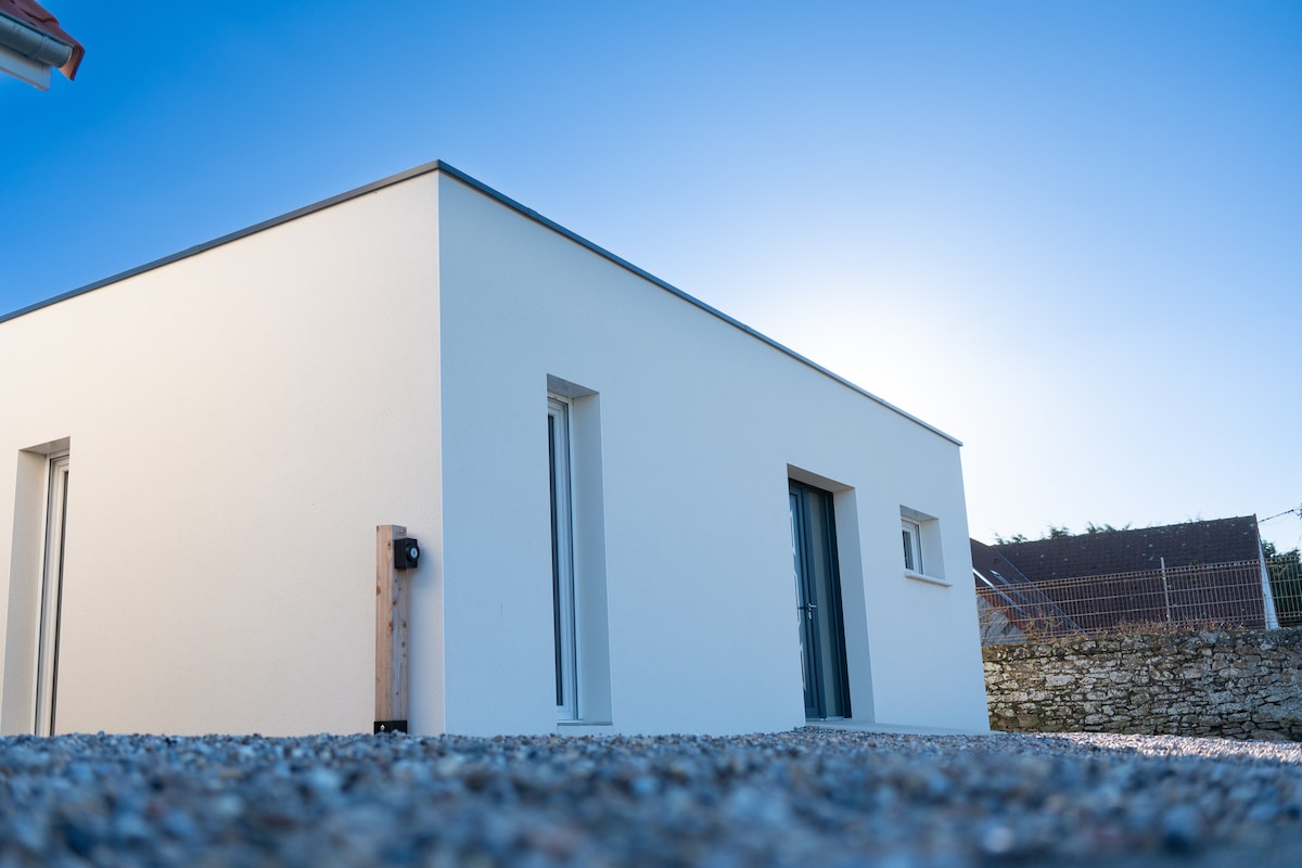 The exterior of the modern building is shown, featuring a smooth, white facade. Large windows allow natural light to enter, while the minimalist design creates a serene appearance. A gravel pathway leads to the entrance, enhancing accessibility for all guests.