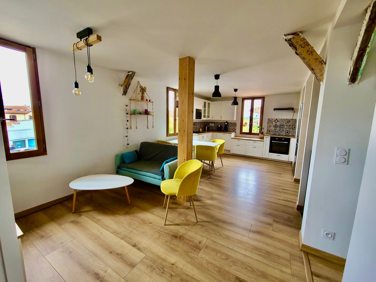 The spacious living area features a green sofa beside a round white coffee table. Yellow chairs are positioned around the dining table, and large windows allow natural light to fill the room. Exposed beams add character to the modern design.