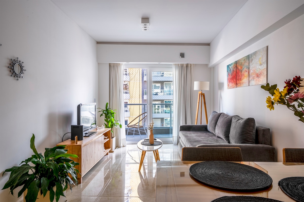 A modern living space features a comfortable grey sofa and a wooden coffee table, surrounded by natural light streaming through wide glass doors. A small potted plant adds a touch of greenery, while a dining area is visible with a set of four chairs arranged around a circular table.