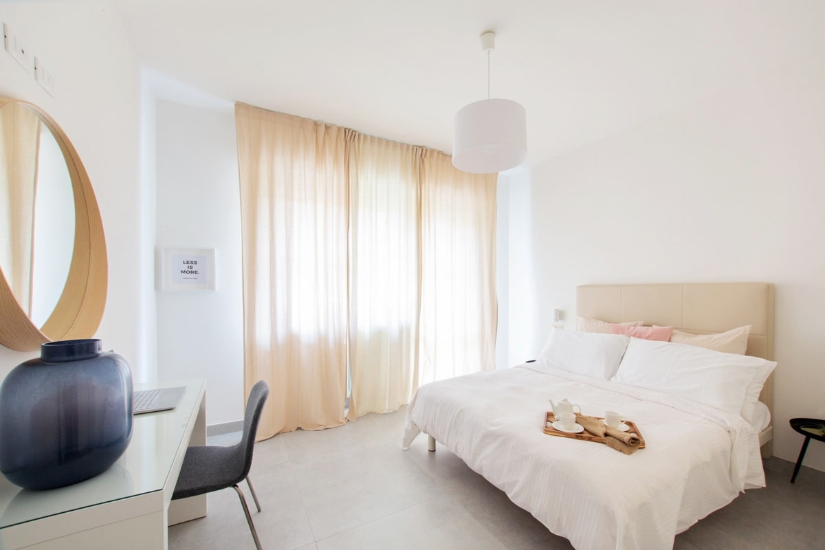 A spacious bedroom is filled with natural light, featuring a large bed with white linens and soft pillows. A minimalist desk and chair are present, along with a round mirror and a decorative dark vase. Light curtains frame the window, enhancing the airy ambiance.