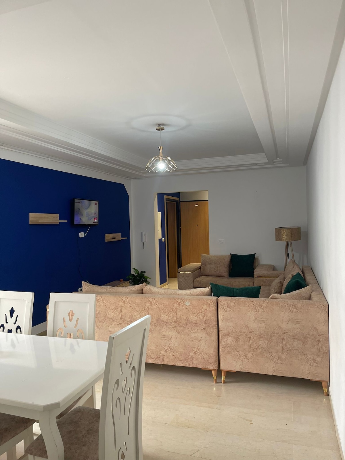 An inviting living area features a light-colored sofa with green pillows, positioned against a deep blue accent wall. A flat-screen television is mounted on the wall, while a dining table with white chairs occupies the foreground, enhancing the space's open and welcoming design.