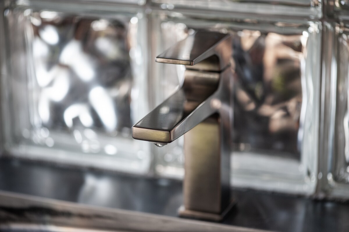 A sleek, modern faucet is positioned over a dark countertop. The faucet features a contemporary design with clean lines, and water is seen starting to drip from the spout. Background glass bricks provide a textured, reflective surface.