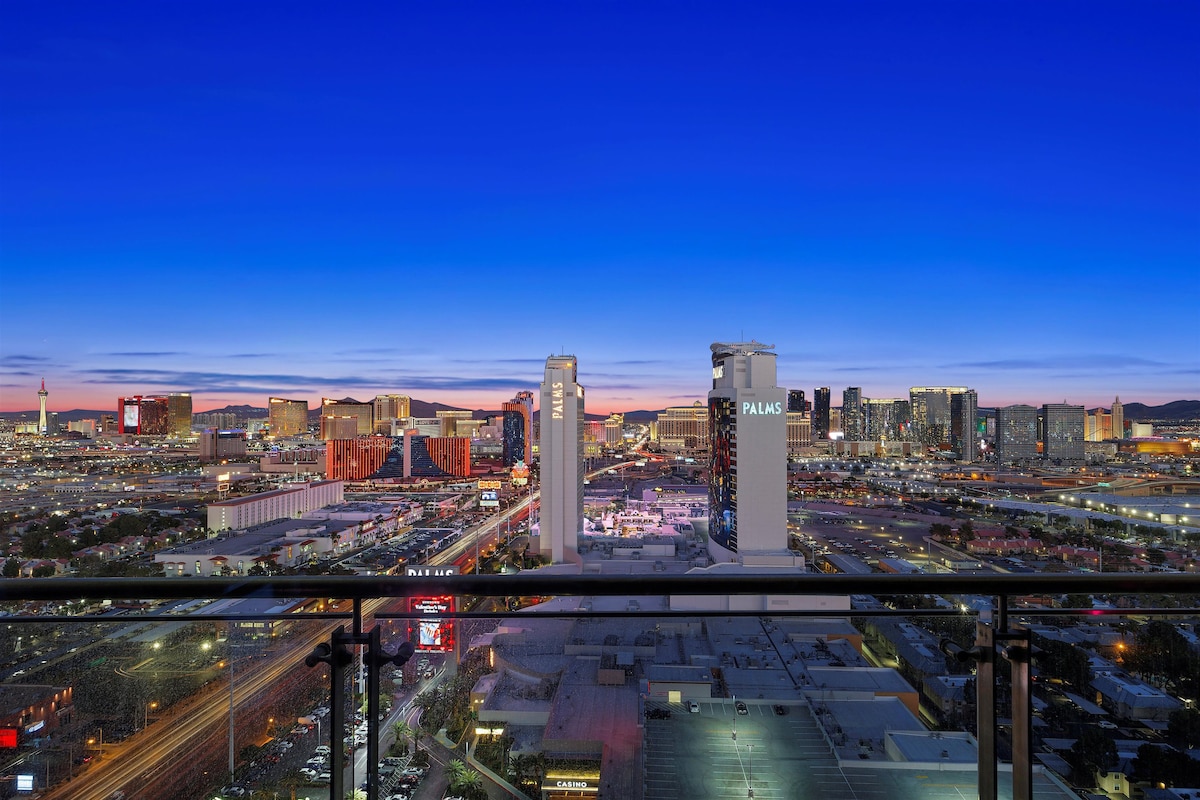 A panoramic view of the Las Vegas Strip is showcased from the balcony, highlighting the skyline bathed in twilight colors. Tall buildings and vibrant lights are visible, creating a dynamic urban landscape against a deep blue and orange sky.