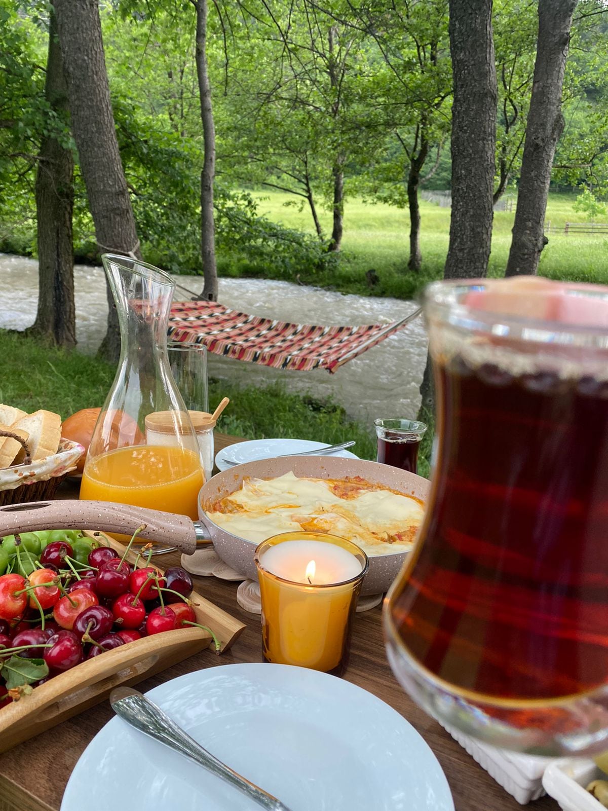 A dining setup features a selection of fresh fruits, bread, and a warm dish, complemented by a glass of juice and a lit candle. A gentle stream flows nearby, surrounded by lush greenery and trees, enhancing the tranquil outdoor setting.
