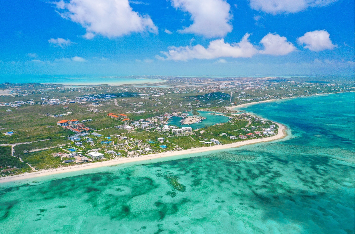 The aerial view captures the vibrant turquoise waters of the Caribbean Sea, contrasting against a sandy shoreline. Lush greenery and colorful buildings dot the landscape, indicating the presence of resorts and residential areas, while the sky features scattered clouds under a bright blue backdrop.