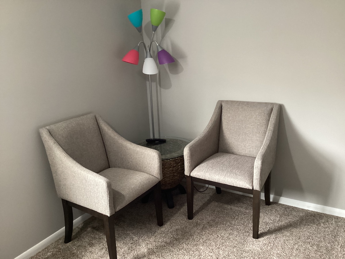 A corner seating area features two upholstered chairs with tapered legs, positioned around a small round table. A multi-colored floor lamp stands nearby, providing additional lighting with its soft hues. The carpeted floor adds a comfortable touch to the space.