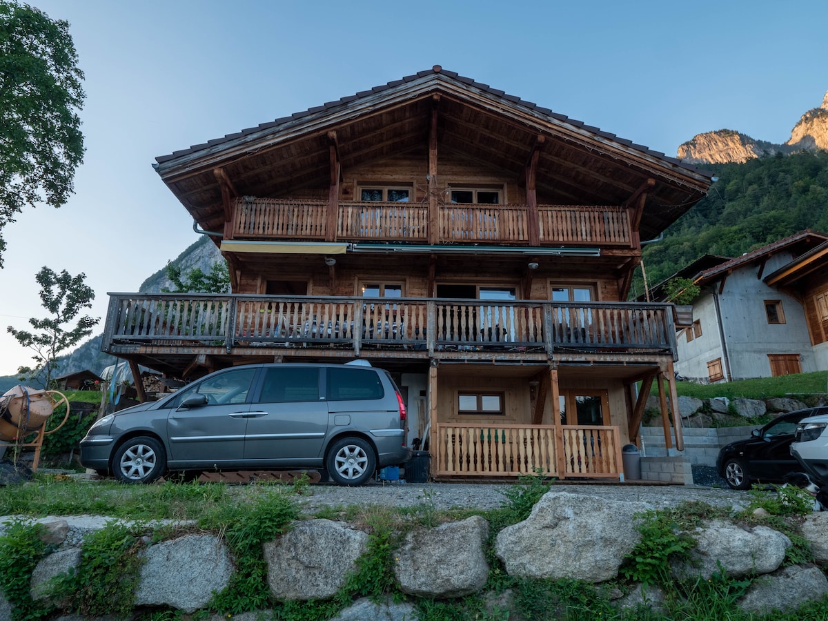 A multi-level wooden chalet is showcased, featuring prominent balconies with wooden railings. A gray vehicle is parked in front on a gravel driveway, surrounded by greenery and large stone landscaping. The chalet is flanked by mountainous terrain, enhancing its scenic setting.
