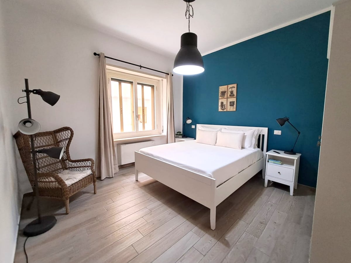 A bright sleeping area features a white bed with a minimalist design, complemented by soft pillows. A cozy chair made of woven material is positioned nearby. Natural light enters through a window dressed in light curtains, while a dark accent wall adds contrast to the space.