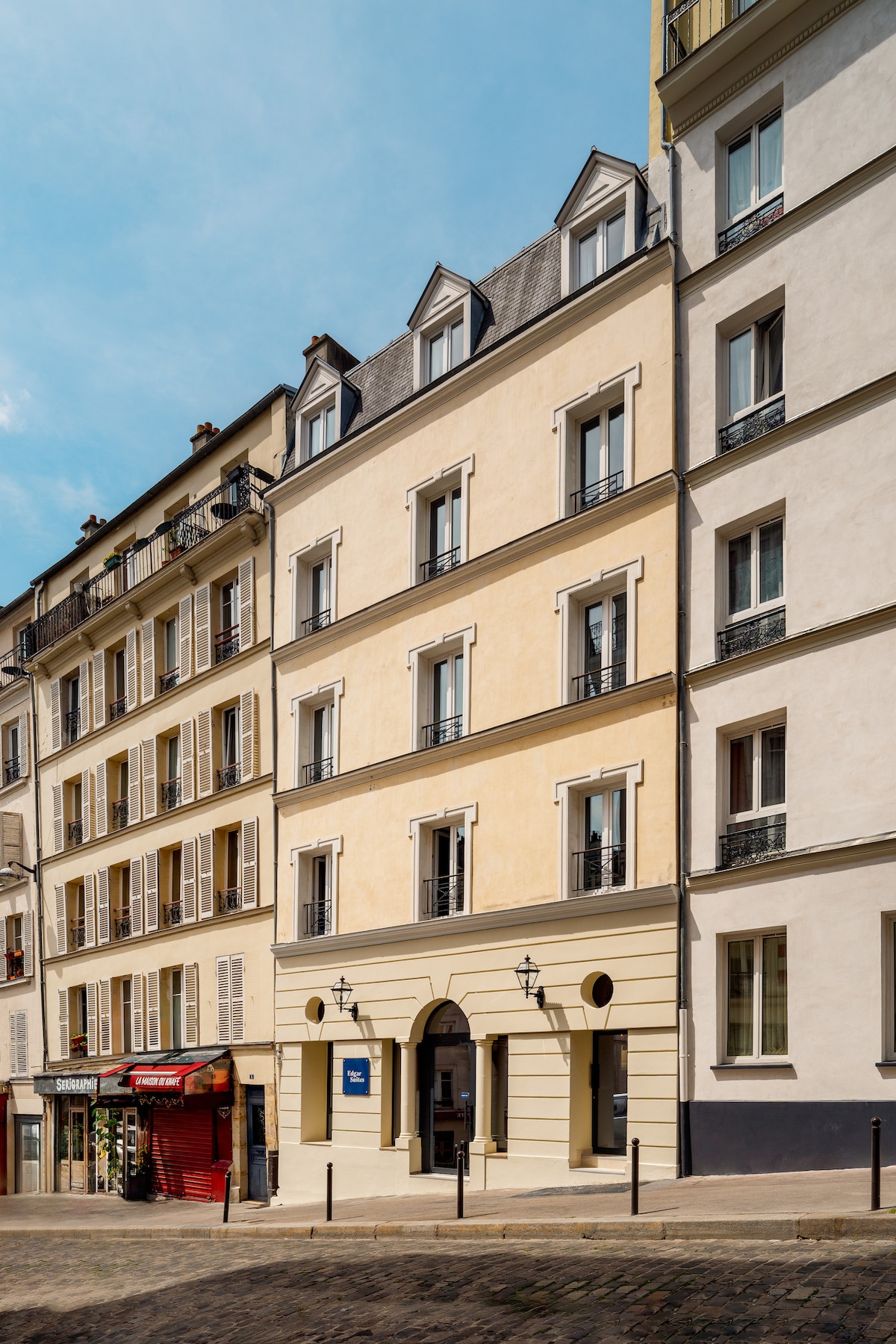 The exterior of the building is showcased, featuring a classic Parisian architectural style with beige and cream-colored facades. The entrance is marked by contemporary light fixtures, and tall windows are accented by black iron railings. Cobbled streets add to the neighborhood's charm.