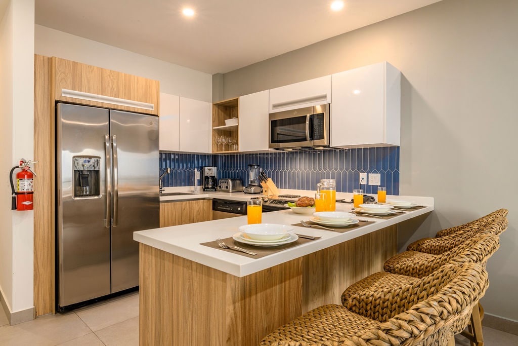 A modern kitchen is presented, featuring stainless steel appliances, including a refrigerator and a microwave. A prominent kitchen island with bar seating is visible, set with plates and drinks. Blue tiled backsplash adds a clean touch to the light-colored cabinetry.