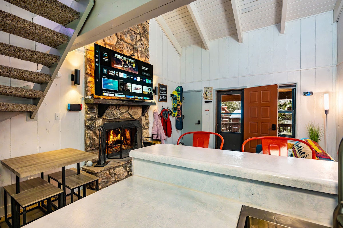 A living area with a stone fireplace and mounted television displays various channels. A dining table with four chairs is positioned nearby, with a well-equipped kitchen counter in the foreground. Stairs lead to the loft above, while views of the outdoors are visible through the open door.
