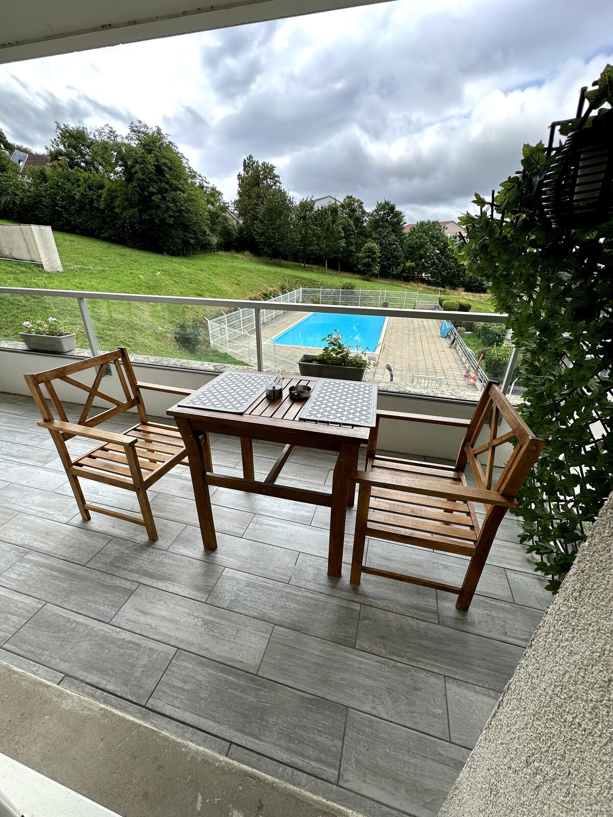 An outdoor balcony features a wooden table with two matching chairs, overlooking a grassy area and a swimming pool. The pool, visible in the background, is surrounded by a secure fence, and greenery frames the balcony view.