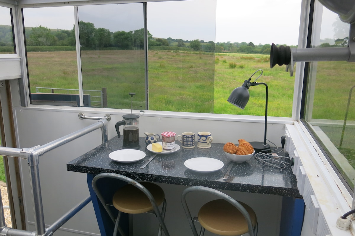 A kitchen area is seen with a black speckled countertop and minimalist seating. Two plates are set with bread and butter, accompanied by croissants and a small container of jam. A lamp casts light on the setup, while a scenic view of the countryside is visible through large windows.