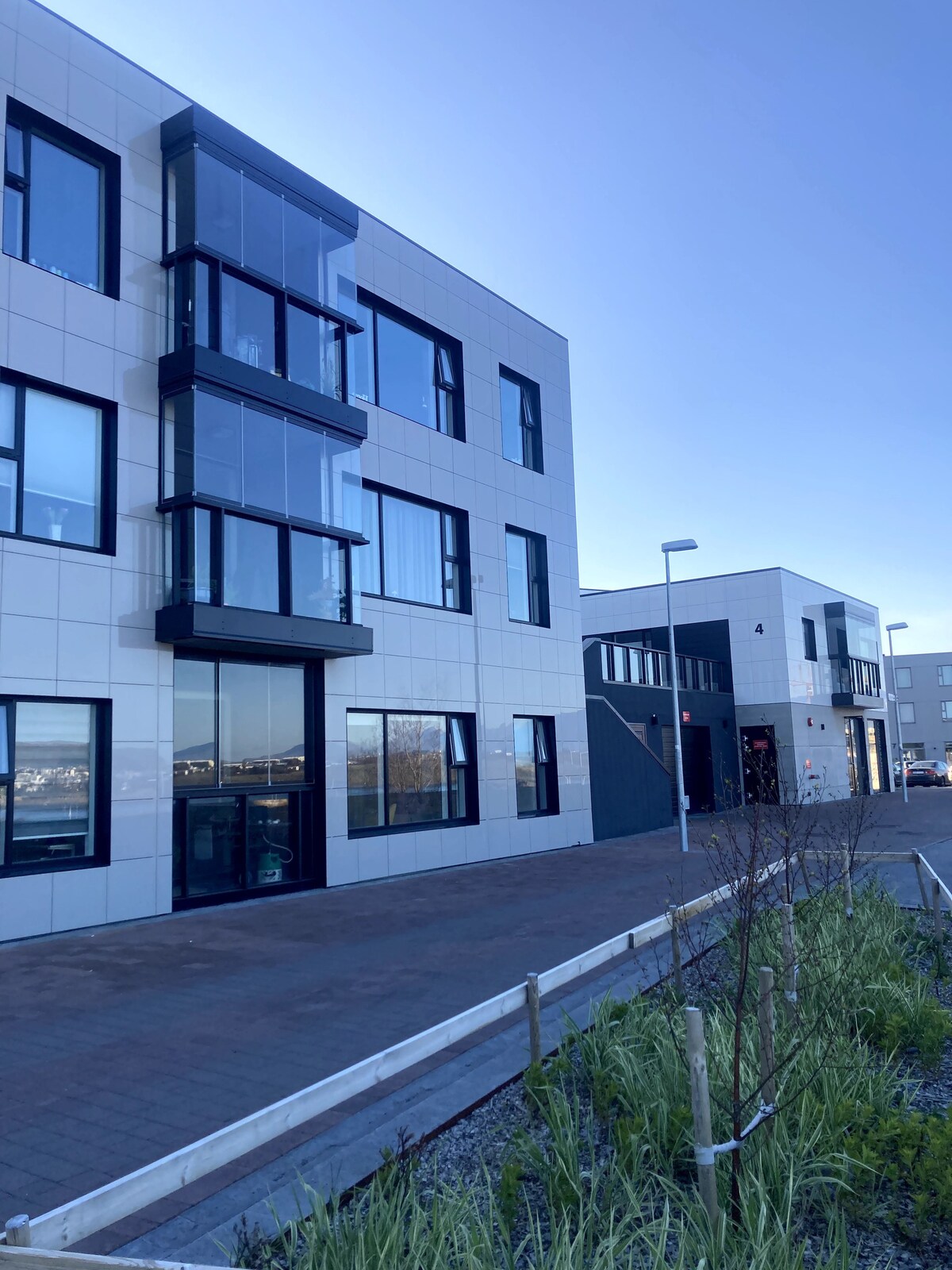 A modern apartment building features sleek architecture with large glass windows. The light-colored facade is complemented by a clean pathway lined with greenery. Nearby structures are visible, indicating a quiet neighborhood setting.