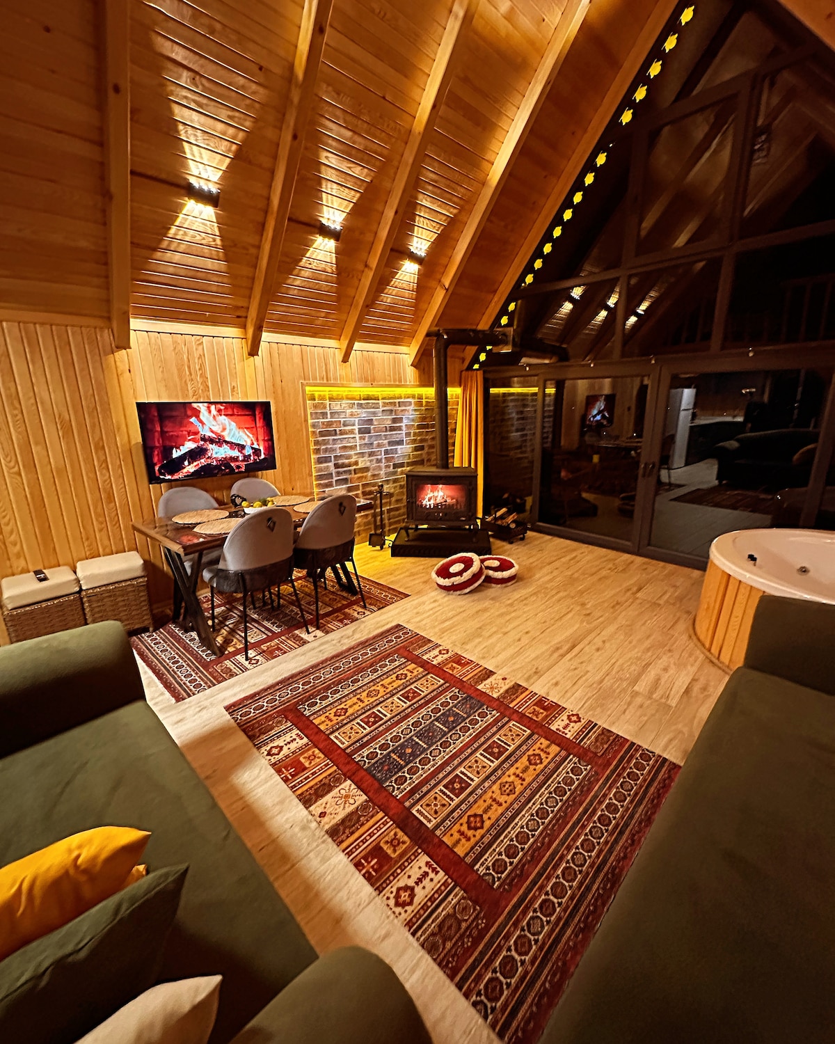 A warm and inviting living area features a wooden ceiling with exposed beams, illuminated by soft lighting. A dining table with four chairs is positioned beside a stylish electric fireplace, creating a cozy ambiance. A Jacuzzi is visible in the background, enhancing relaxation options.