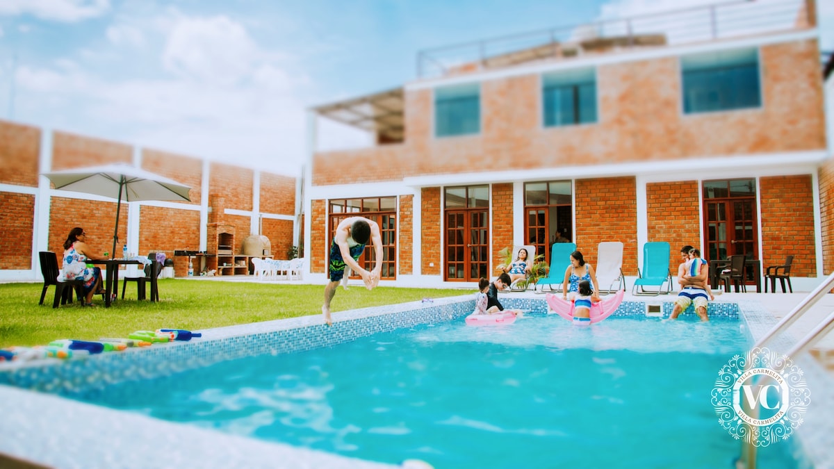 A refreshing swimming pool is surrounded by lush green grass and outdoor furnishings, including lounge chairs and an umbrella-shaded table. Several guests are enjoying the pool, with one person captured in mid-air above the water, creating a lively atmosphere.