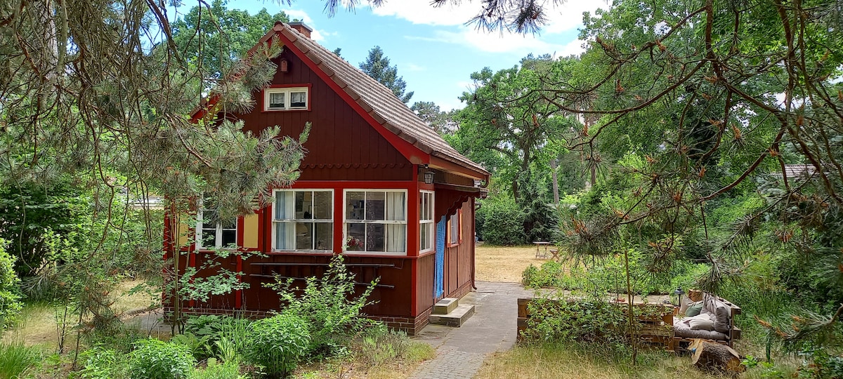 A charming summer house from the 1930s is framed by lush greenery. The red wooden exterior features white windows and a welcoming entrance. Surrounding trees provide shade, and a well-maintained garden space is visible, creating a serene outdoor environment.