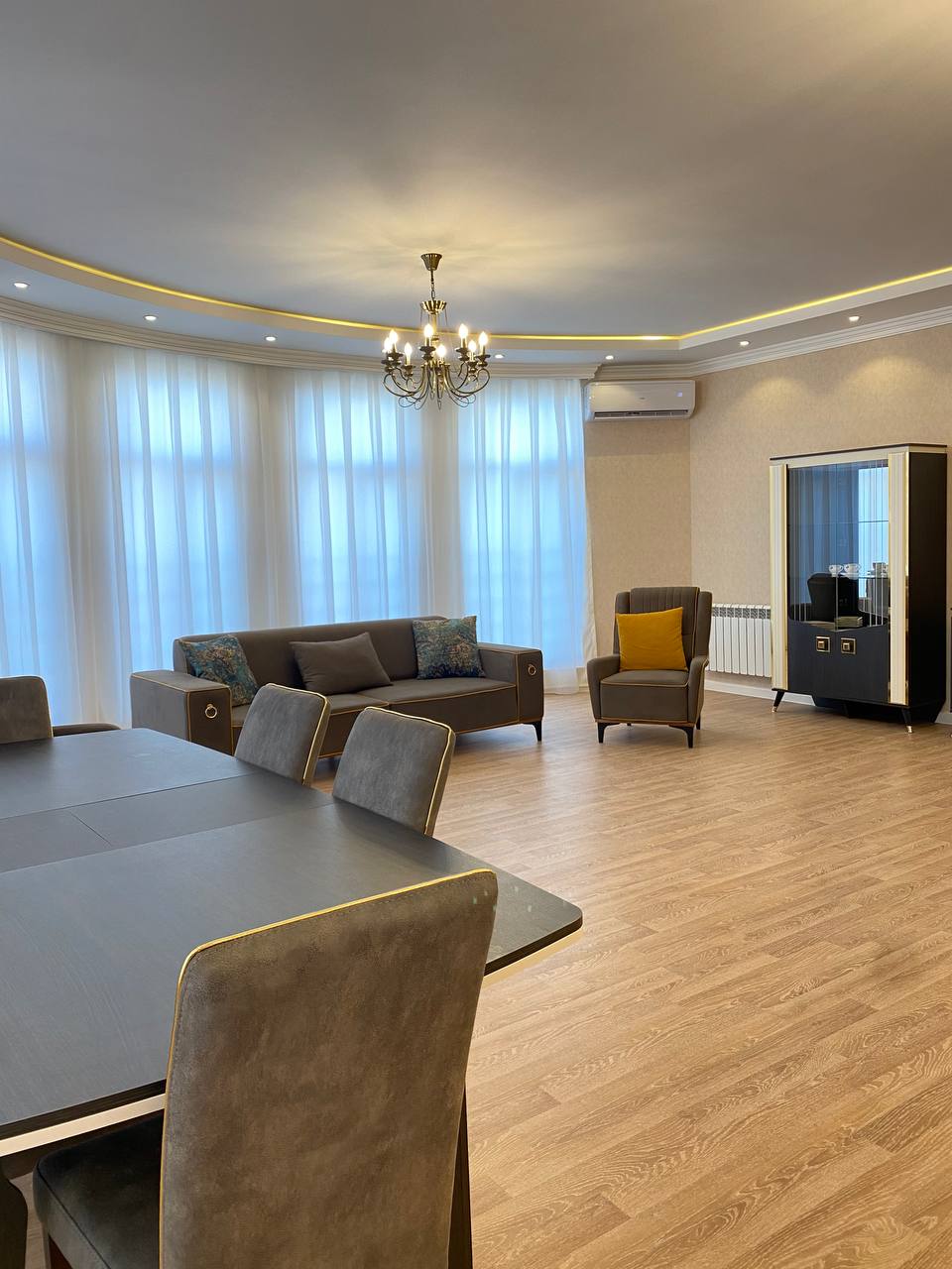 A spacious living area features large windows draped with sheer white curtains, allowing ample natural light. A sleek dining table with upholstered chairs sits adjacent to a modern sofa and a stylish armchair. Elegant lighting is provided by a central chandelier.