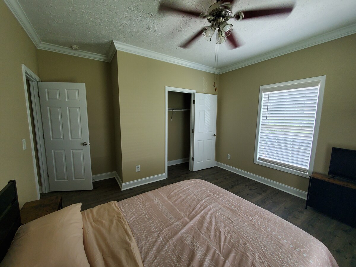 A well-lit bedroom features a spacious bed covered with a soft beige comforter. The walls are painted in a warm light hue, and a ceiling fan is visible above. A window allows natural light to flow in, complemented by a closet and doorway.