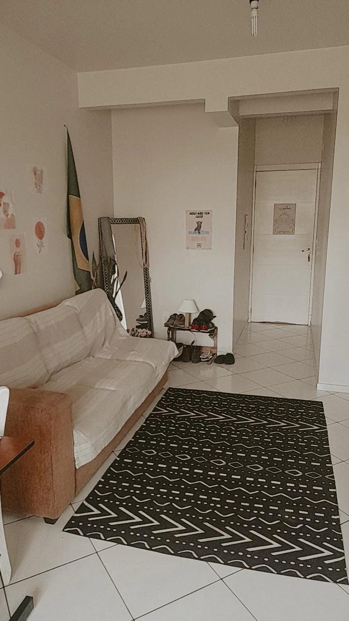 A cozy living area features a light-colored sofa against a neutral wall. A patterned rug is positioned on the tiled floor, and a wall-mounted mirror reflects part of the room. The entrance to the apartment is visible in the background, framed by a simple door.