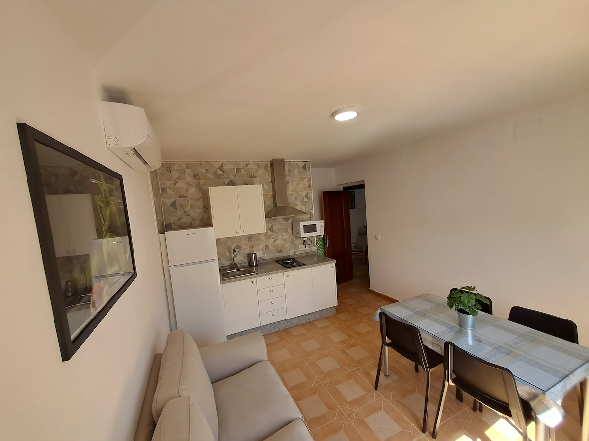 The open-plan living area is highlighted by a neutral palette and features a soft sofa, a dining table with four chairs, and a compact kitchen with modern appliances. Natural light filters through the space, enhancing the clean lines and functional design.