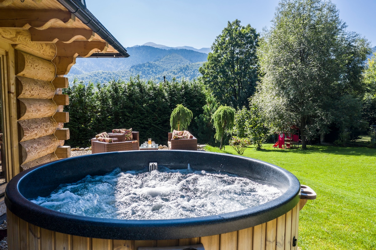 A hot tub is prominently featured on a wooden deck, with water bubbling invitingly. In the background, lush green trees and mountains are visible under a clear blue sky. Comfortable seating areas are arranged nearby, enhancing the outdoor space.