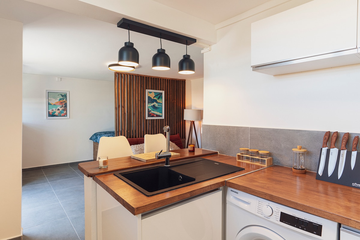 A modern kitchen area features sleek cabinetry and wooden countertops. Three pendant lights hang above a deep sink. Wooden accents add warmth, with a dining table placed nearby. In the background, a cozy sleeping area is visible, separated by wooden slats, enhancing the open layout.