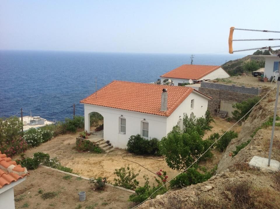 A white house with a terracotta roof is positioned overlooking the Mediterranean Sea, surrounded by natural landscaping. Shrubs and flowers in varying colors create a serene environment. The property is set on a hillside with additional buildings visible in the background.