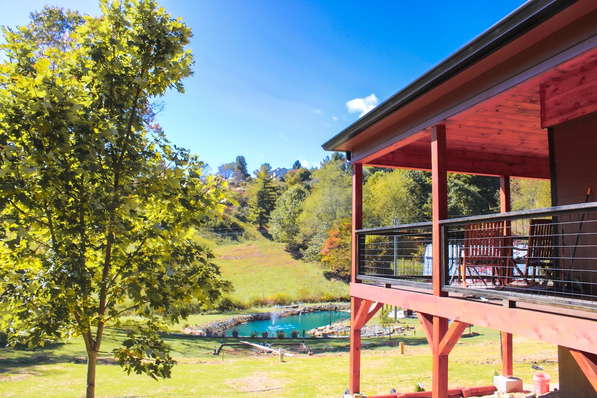 A porch with a wooden railing offers a view of a tranquil pond surrounded by lush greenery. Sunlight filters through the leaves of a nearby tree, enhancing the serene environment. Rolling hills are visible in the background, creating a peaceful outdoor setting.
