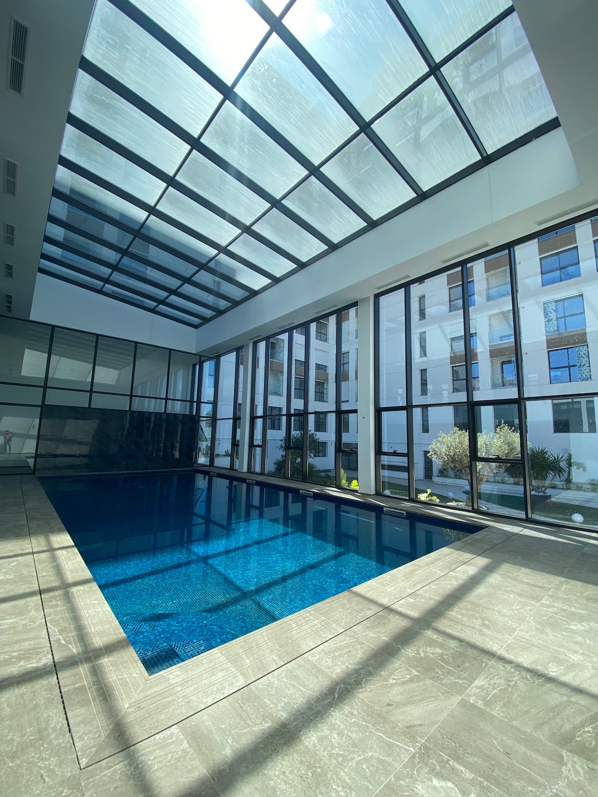 An indoor pool area is highlighted by a glass ceiling, allowing natural light to fill the space. Large windows surround the pool, providing views of the exterior greenery. The pool is surrounded by a stone tile floor, creating a serene and modern atmosphere.