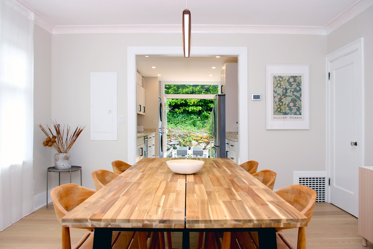 A spacious dining area features a wooden table surrounded by several chairs, emphasizing a modern and minimalistic design. Natural light enters through an open doorway that leads to an outdoor patio area, creating a seamless connection between indoor and outdoor spaces.
