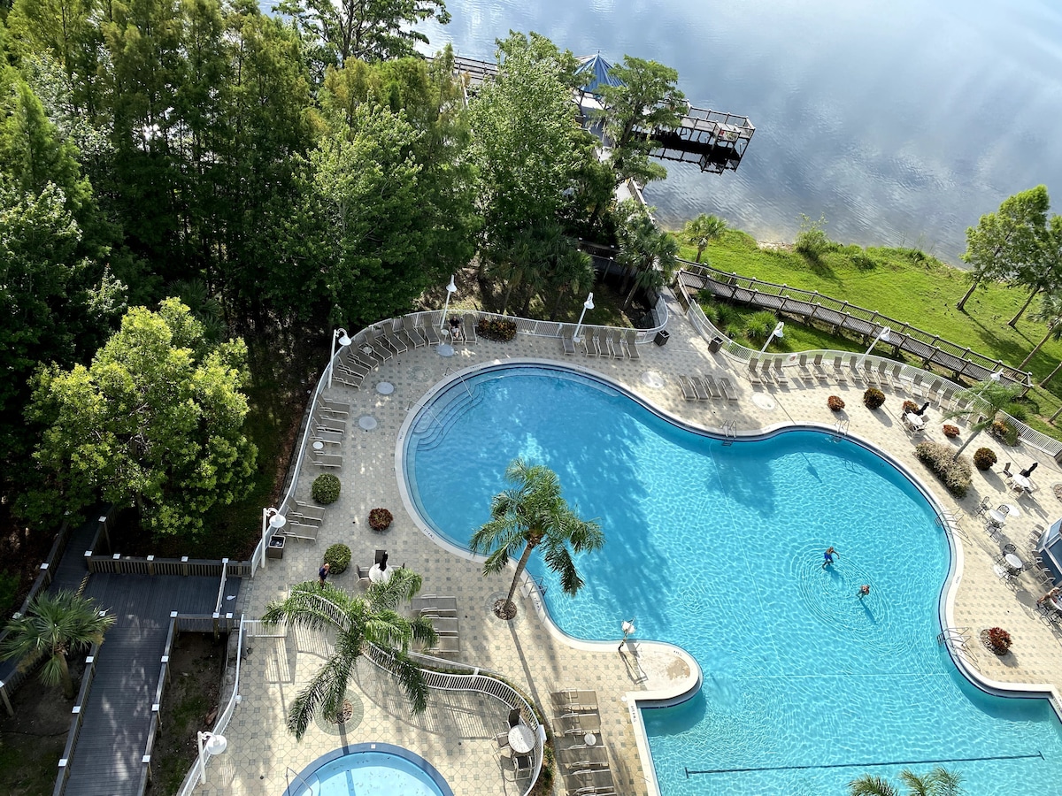 An aerial view captures a large swimming pool curving gracefully, surrounded by palm trees and lounge chairs. Adjacent to the pool, a kiddie pool is visible. The tranquil waters of Lake Bryan glimmer in the background, bordered by lush greenery.