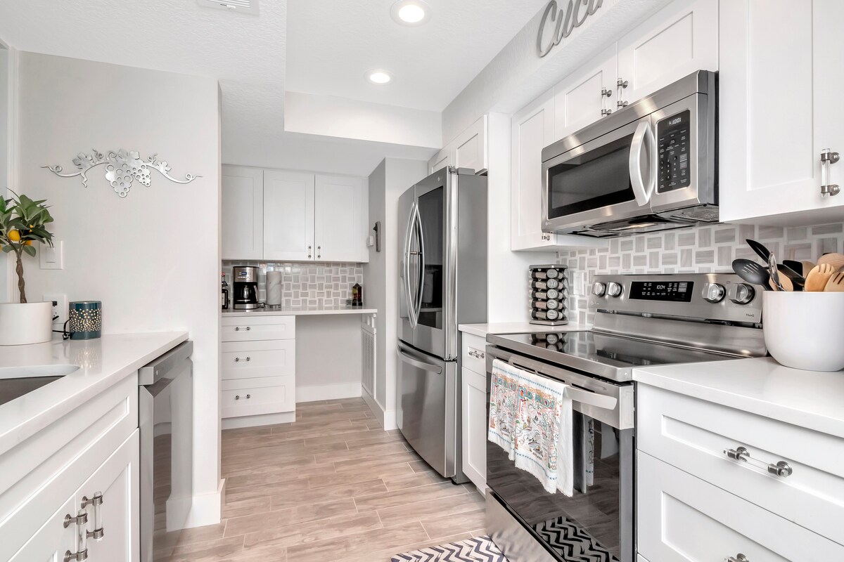 The modern kitchen features white cabinetry, stainless steel appliances, and a stylish backsplash. A double sink and ample counter space are complemented by a coffee station in the corner. Natural light fills the room, creating a bright and functional cooking area.