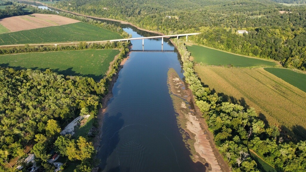 An aerial view captures the serene Osage River meandering through lush green landscapes. Fields of crops line the banks, and a bridge crosses the river, providing a sense of connection to the natural surroundings. The scenery reflects the tranquility of the area.