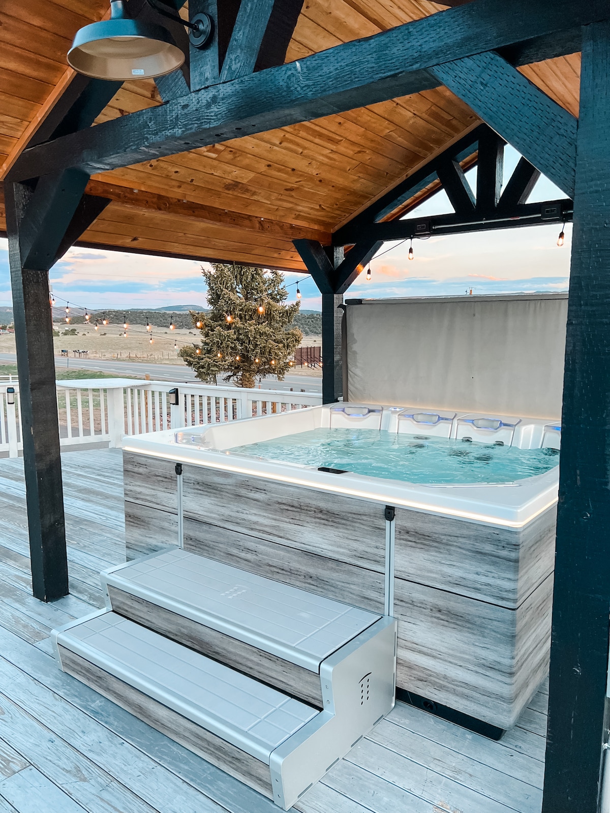 A hot tub is situated under a wooden pergola, with a light wooden deck surrounding it. The area features steps for easy access, and string lights hang above, adding to the ambiance. Scenic views of the valley and distant mountains are visible in the background.
