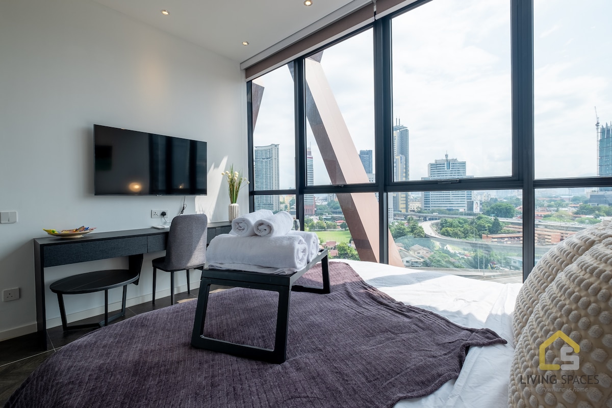 A spacious room features large glass windows that allow ample natural light and provide a clear view of the cityscape. A neatly arranged bed with a folded towel sits at the center, complemented by a small table and two chairs for added convenience.